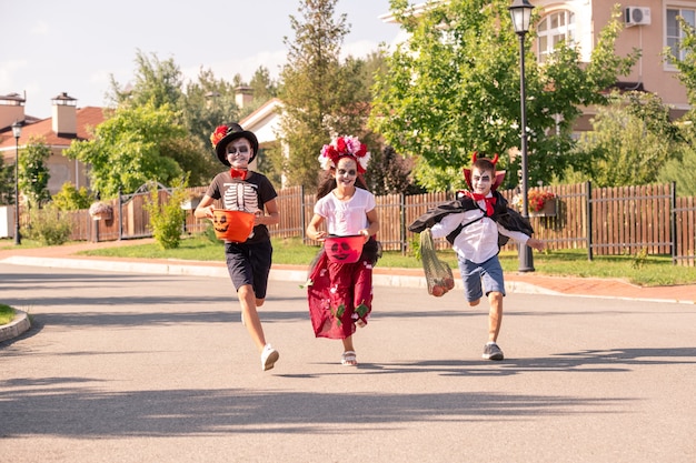 Drei freudige Kinder in Halloween-Kostümen, die Behälter mit süßen Leckereien halten, während sie an sonnigem Herbsttag die breite Straße hinunterlaufen