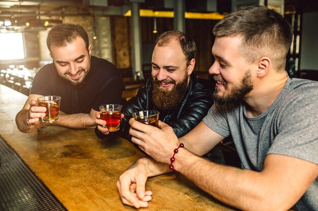Drei Freunde sitzen zusammen an der Theke. Sie halten Gläser Alkohol in den Händen. Männer lächeln.