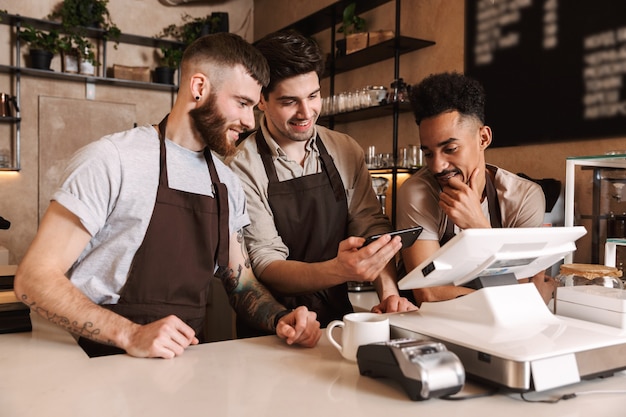 Drei fröhliche Baristas stehen hinter der Theke im Café und benutzen Handy