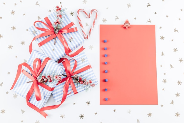 Drei Geschenkboxen in einem blauen Streifen mit roten Bändern, Süßigkeiten, Weihnachtsbäumen aus Holz, einem flauschigen Schneeball und einem leeren Blatt Papier auf einem weißen