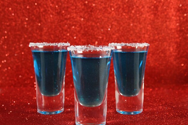 Drei Gläser mit blauen alkoholischen Getränken stehen auf einem rot glänzenden Hintergrund