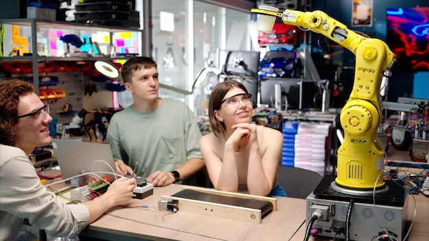 Drei glückliche junge Ingenieure reparieren einen gelben Roboterarm in der Werkstatt Computerprogrammierung