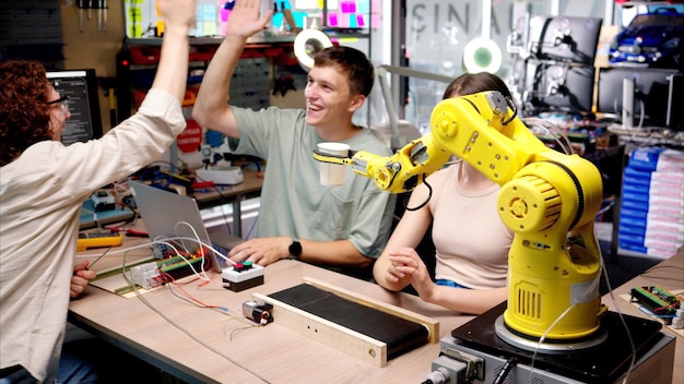 Drei glückliche junge Ingenieure reparieren einen gelben Roboterarm in der Werkstatt Computerprogrammierung