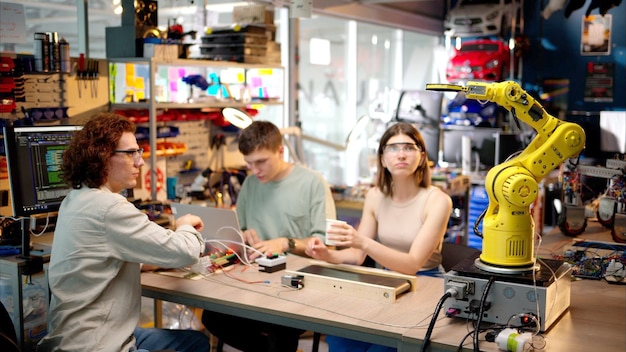 Drei glückliche junge Ingenieure reparieren einen gelben Roboterarm in der Werkstatt Computerprogrammierung