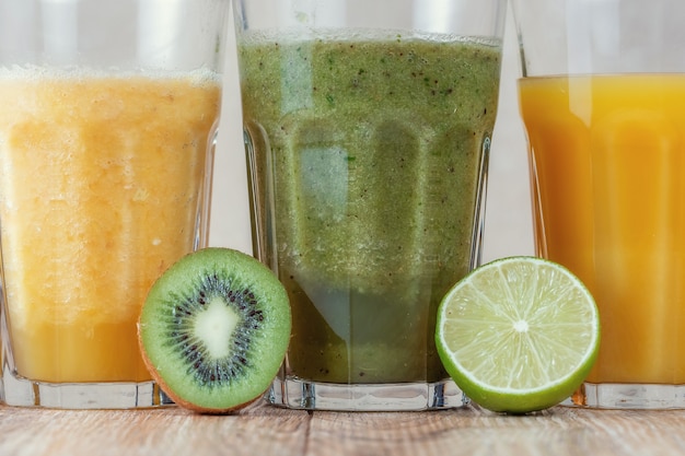 Drei hohe Gläser mit Kiwi- und Spinat-Smoothies in einer Reihe, umgeben von Früchten auf einem Holztisch.