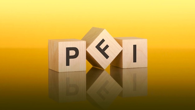 Drei Holzwürfel mit den Buchstaben PFI auf der leuchtend gelben Oberfläche, die Aufschrift auf den Würfeln spiegelt sich in der grauen Oberfläche des Tisches wider