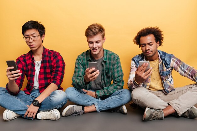 Drei hübsche junge Männer Freunde mit Handy über gelbem Hintergrund