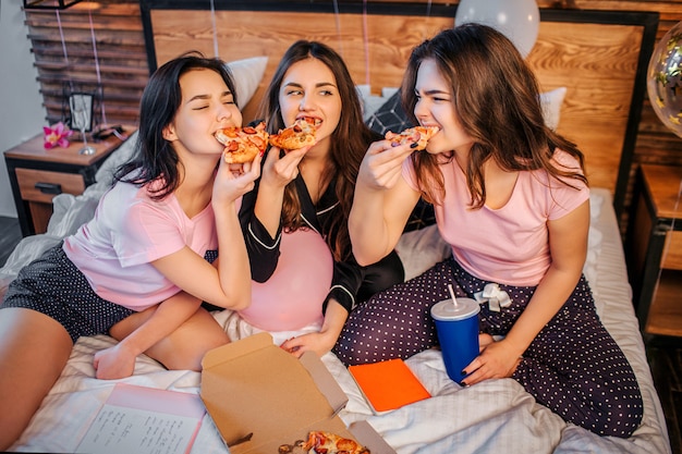 Drei hungrige Jugendliche, die Stücke von der Pizza beißen. Sie verschlingen es. Hungrige Frauen tragen Pyjamas. Sie sitzen auf dem Bett im Zimmer.