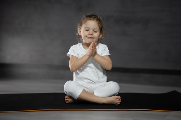 Drei Jahre altes kleines Mädchen, das in einer Lotus-Pose auf einem grauen Hintergrund in einem dunklen Raum meditiert