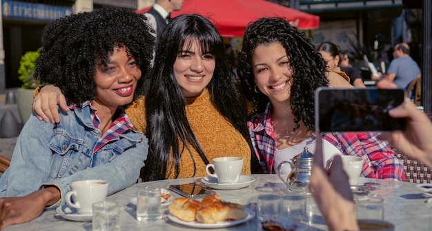 Drei junge Frauen, eine haitianische Afro-Frau und zwei Latinas lächeln, als sie sich in einem Straßencafé fotografieren lassen