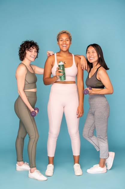Drei junge lachende Sportlerinnen in Activewear, die eine Flasche Wasser und Hanteln halten, während sie zusammen trainieren