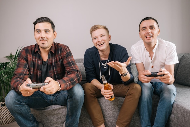 Drei junge Männer mit Joysticks spielen Fußball im Fernsehen. Guy hat in einem Konsolen-Videospiel gegen seinen Freund verloren. Guy hält eine Flasche Bier in seinen Händen