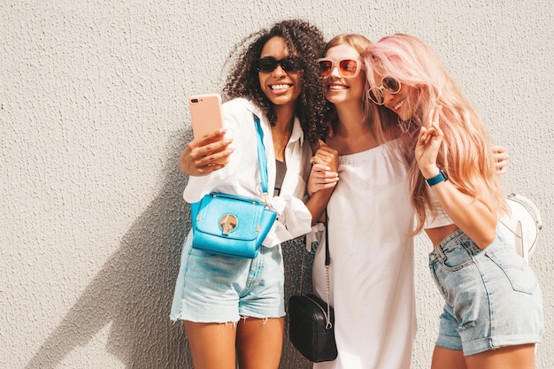 Drei junge schöne lächelnde Hipster-Frauen in trendiger SommerkleidungSexy sorglose gemischtrassige Frauen posieren auf dem StraßenhintergrundPositive Models, die Spaß mit Sonnenbrillen haben Selfie-Fotos machen