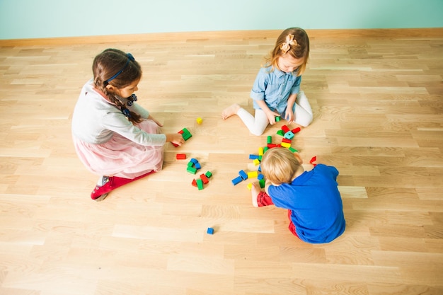 Drei Kinder spielen auf einem Holzboden mit mehrfarbigen Blöcken in ihrem Zimmer, Draufsicht. Kinder unterschiedlichen Alters spielen allein im leeren Raum