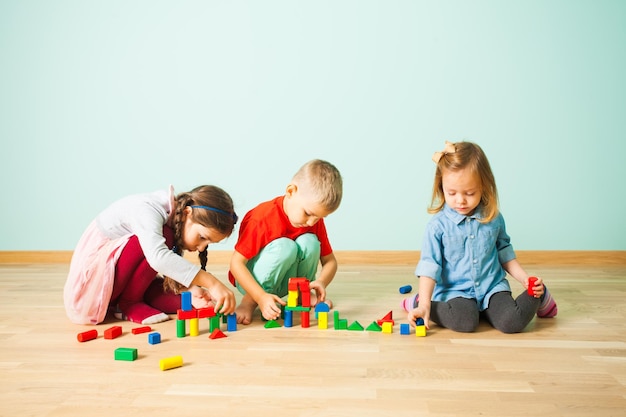 Drei kleine Freunde, hübscher Junge und süße Mädchen, die zu Hause spielen und auf dem Holzboden sitzen und Burgen aus bunten Holzklötzen bauen. Kreative Kinder verbringen ihre Freizeit damit, mit Bauklötzen zu spielen