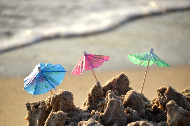Drei kleine Sonnenschirme aus Papier für Cocktails stehen im Sand am Sandstrand von Meer oder Ozean und rollenden Wellen an sonnigen Tagen in Nahaufnahme. Konzept Glück Freude entspannen Tourismus Urlaub Reisen