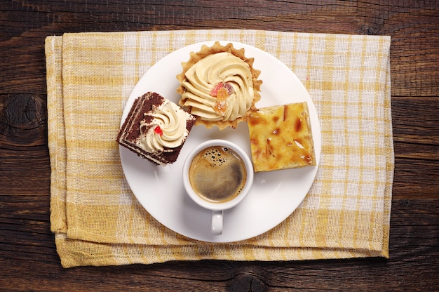 Drei Kuchen und Kaffeetasse im weißen Teller auf einer gelben Tischdecke. Ansicht von oben