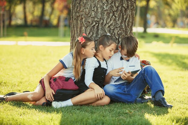 Drei lächelnde Teenager-Schulfreunde, die im Park auf Gras sitzen und neues Tablettenspiel zusammen spielen. Unterschiedliche Emotionen in ihren Gesichtern