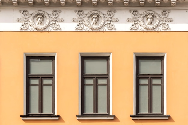 Drei leere Fenster mit dunkelbraunen Rahmen an der Fassade eines gelben Hauses mit historischen Ornamenten