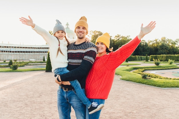 Drei Leute haben eine gute Zeit zusammen, Mutter, Vater und kleine Tochter stehen nebeneinander, gehen im Freien, winken mit den Händen, haben fröhliche Gesichtsausdrücke. Menschen, Gesichtsausdrücke und Emotionen