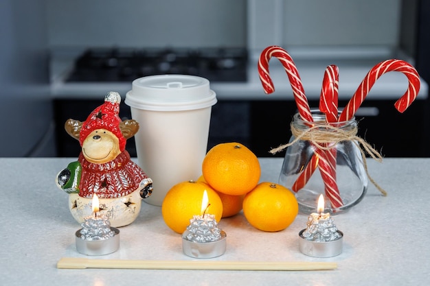 Drei Mandarinen und Essstäbchen, eine Weihnachtsfigur und Süßigkeiten auf einem weißen Tisch vor dunklem Küchenhintergrund