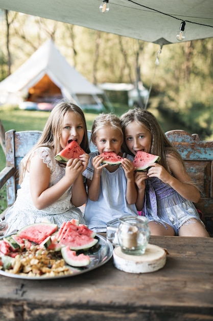 Drei niedliche glückliche lächelnde Mädchen, Schwestern, Unholde, die am Tisch auf Weinlese-Holzbank sitzen und Wassermelone draußen essen