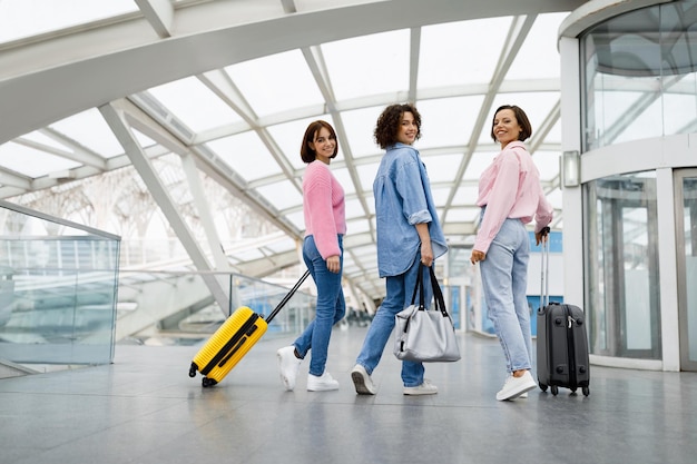 Drei schöne Frauen, die mit Gepäck am Flughafen spazieren gehen und sich in die Kamera drehen