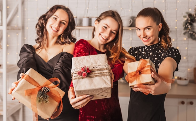 Drei schöne fröhliche glückliche junge Mädchen, die mit Geschenk am Weihnachtsesszimmertisch auf dem Hintergrund der Neujahrsküche lachen