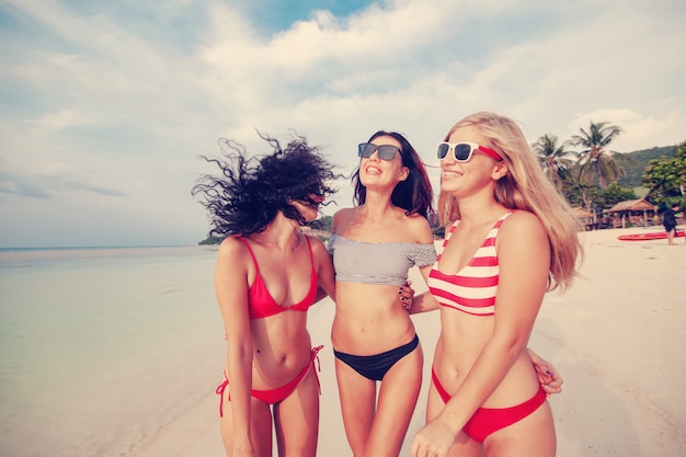 Drei schöne junge europäische schlanke Freundinnen in leuchtend roten und gestreiften Bikinis spazieren an einem tropischen Strand im Urlaub, Glück, Freude, Sommer und Spaß