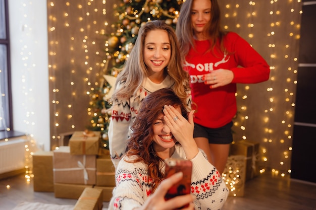 Drei schöne Mädchen machen Selfie am Telefon, Grimasse. Beste Freunde der jungen Frau, die Weihnachten im Haus feiern. Wunderschöner goldener Weihnachtsschmuck an einem hohen Weihnachtsbaum