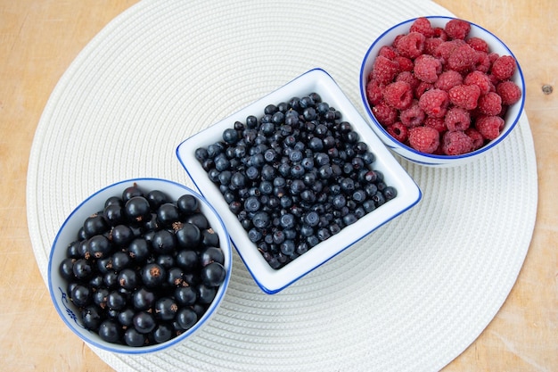 Drei Schüsseln mit Beeren der Saison, eine Mischung aus frischen Beeren auf einer weißen runden Serviette auf einem Holztisch, Blaubeeren, Johannisbeeren, Blaubeeren, Himbeeren