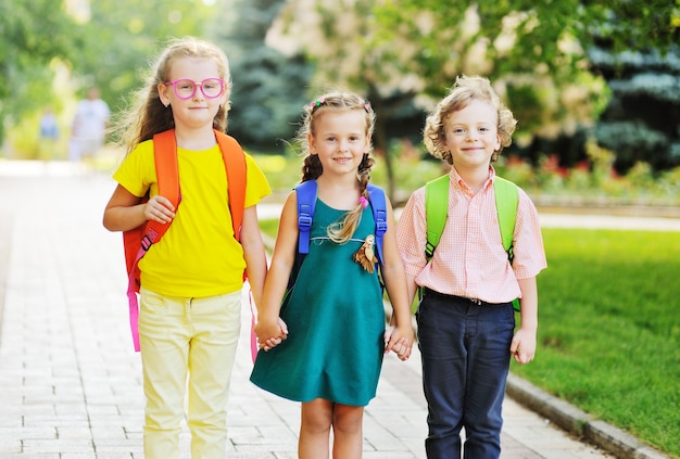 Drei Schulkinder - zwei Mädchen und ein Junge mit lockigen Haaren mit schulfarbenen Taschen lächelnd halten sich an den Händen und gehen zur Schule. Zurück zum Schulkonzept, 1. September, Tag des Wissens, Bildung.
