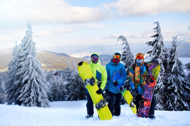 Drei Snowboarder posieren im Skigebiet Freunde, die auf den Berggipfel klettern und ihre Snowboards durch den Wald für Backcountry-Freeride tragen und reflektierende Schutzbrillen tragen, bunte Modekleidung