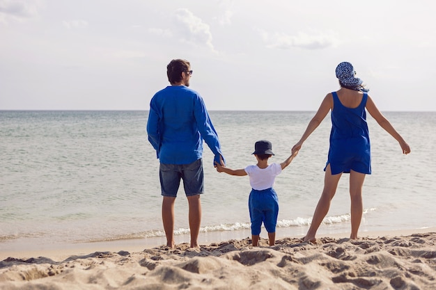 Dreiköpfige Familie in blauer Kleidung steht im Sommer im Urlaub an einem Sandstrand am Meer