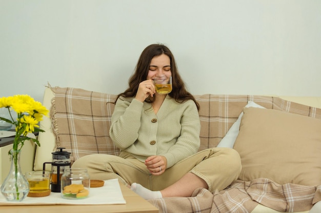 Drinnen in einem hellen Interieur sitzt eine junge fröhliche Frau lächelnd auf einem Sofa und trinkt Kräutertee