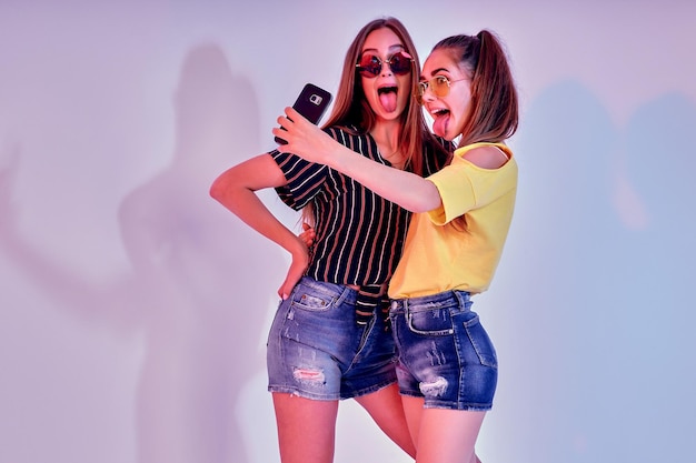 Duas adolescentes em roupas de verão em pé no estúdio e fazendo selfie em fundo branco misturado l...