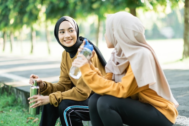 Duas adolescentes sentadas bebendo água com garrafa depois de praticar esportes ao ar livre juntas no campo do jardim