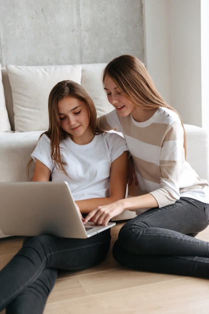 Duas adoráveis jovens adolescentes alegres usando um laptop enquanto estão sentadas em casa