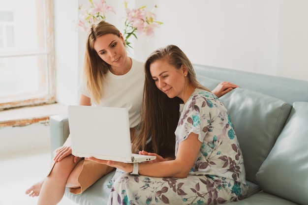 Duas adoráveis jovens conversando em casa no sofá, bebendo café e trabalhando em um laptop