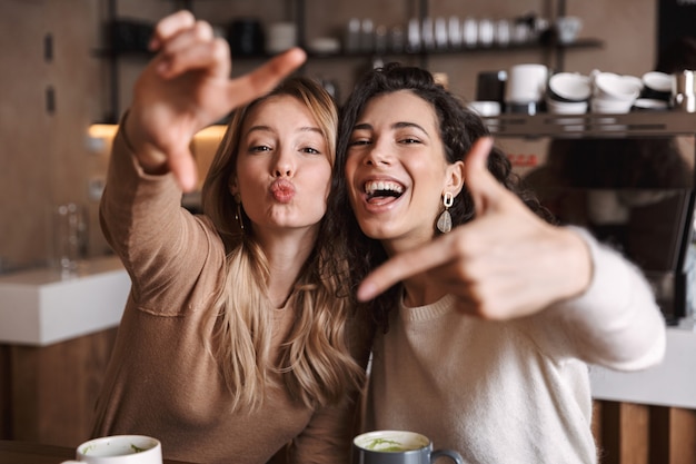 Duas alegres amigas jovens sentadas à mesa do café, se divertindo juntas, bebendo café