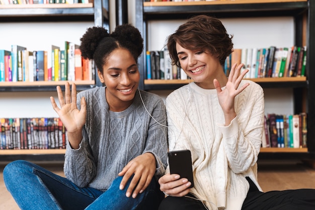 Duas alegres jovens estudantes estudando na biblioteca, ouvindo música com fones de ouvido, segurando um telefone celular