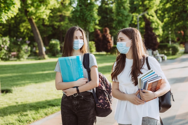 Duas alunas com máscaras médicas protetoras andam e conversam no campus. Educação a Distância. Foco seletivo suave.
