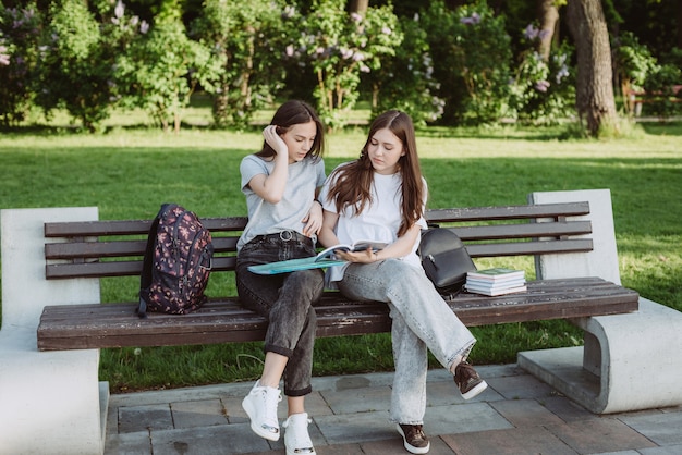 Duas alunas estão olhando para um livro aberto em um banco do parque. Educação a distância, preparação para exames. Foco seletivo suave.