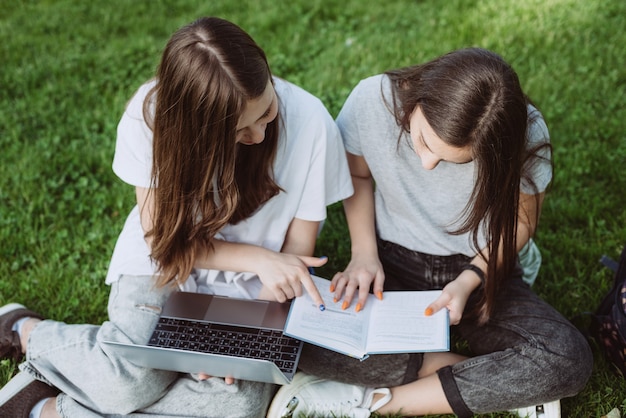 Duas alunas estão sentadas no parque na grama com livros e laptops, estudando e se preparando para os exames. Educação a Distância. Foco seletivo suave.