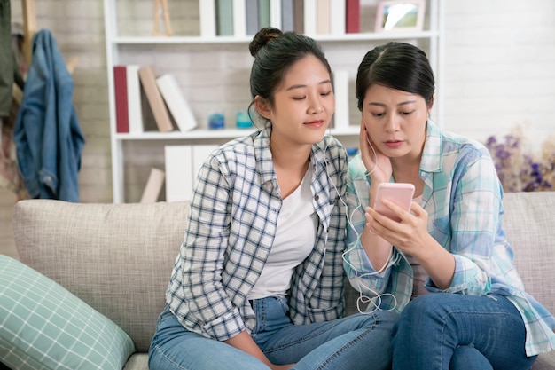 duas amigas adolescentes ouvindo música no sofá na sala de estar em casa. belas mulheres jovens compartilhando fones de ouvido desfrutam de melodia online nova lista de músicas populares olhando juntos na tela do celular.