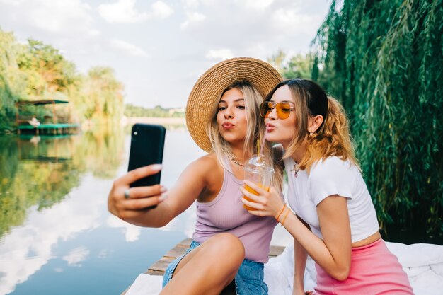 Duas amigas alegres descansando ao ar livre no cais do lago, tirando foto, selfie