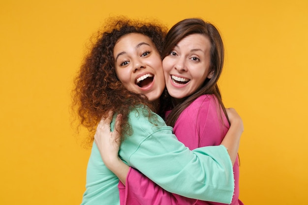 Foto duas amigas alegres e engraçadas garotas europeias e afro-americanas em roupas verdes rosa posando isoladas em fundo amarelo. conceito de estilo de vida de pessoas. mock up espaço de cópia. abraçando, olhando a câmera.