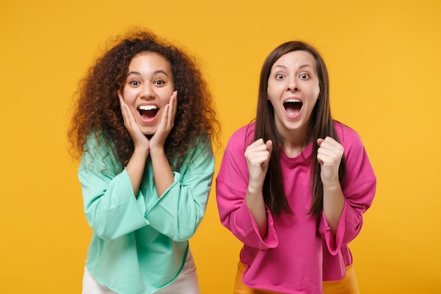 Foto duas amigas animadas garotas afro-americanas europeias em roupas verdes rosa posando isoladas em fundo amarelo. conceito de estilo de vida de pessoas. mock up espaço de cópia. os punhos cerrados colocam as mãos nas bochechas.