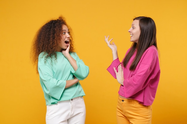 Duas amigas animadas garotas europeias e afro-americanas em roupas verdes rosa posando isoladas em fundo amarelo. Conceito de estilo de vida de pessoas. Mock up espaço de cópia. Olhando um para o outro falando.