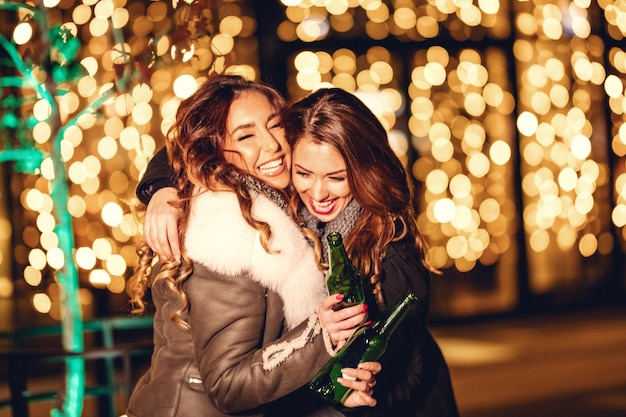 Duas amigas aproveitam a noite abraçando e rindo com garrafas de cerveja nas mãos.